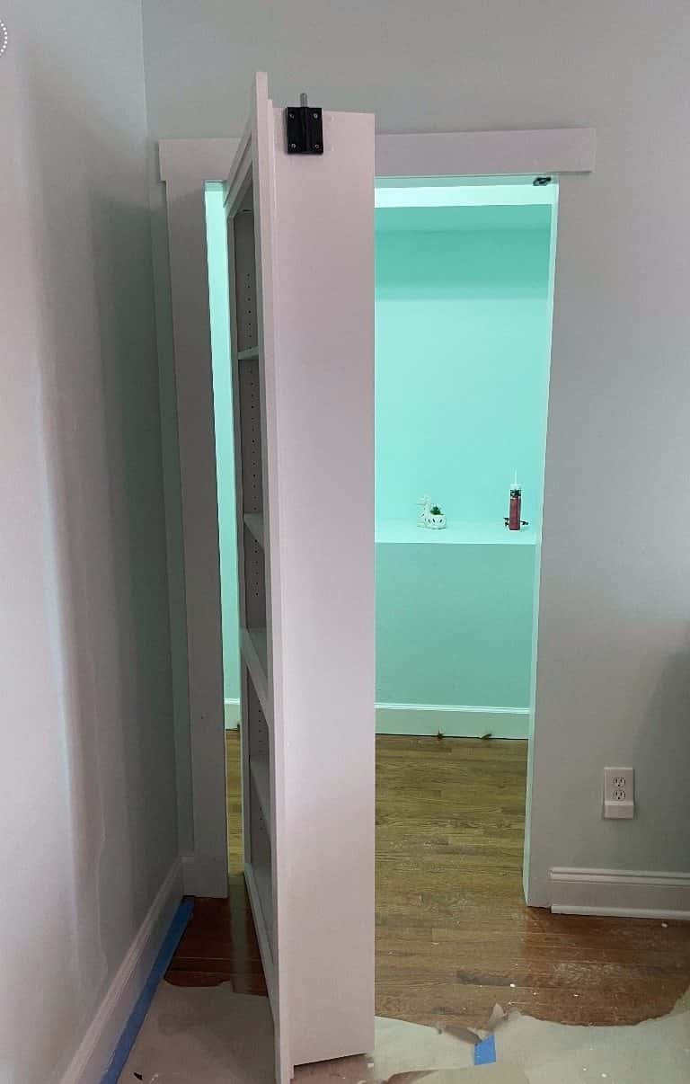 Custom white bookcase hidden door swung open to reveal the room behind it, before final floor and paint touch-up