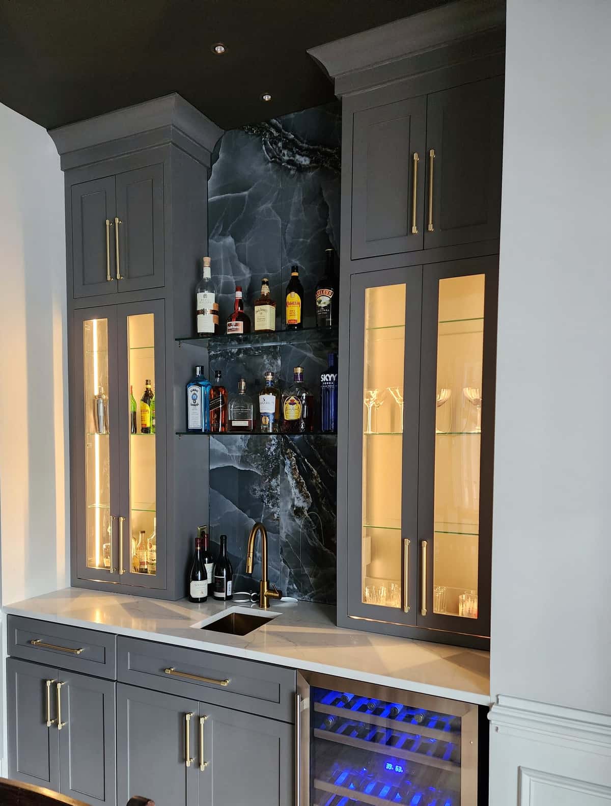 Custom charcoal wet bar with lit glass-front cabinets, black onyx slab backsplash, floating glass shelves, brushed brass hardware and a built-in wine refrigerator
