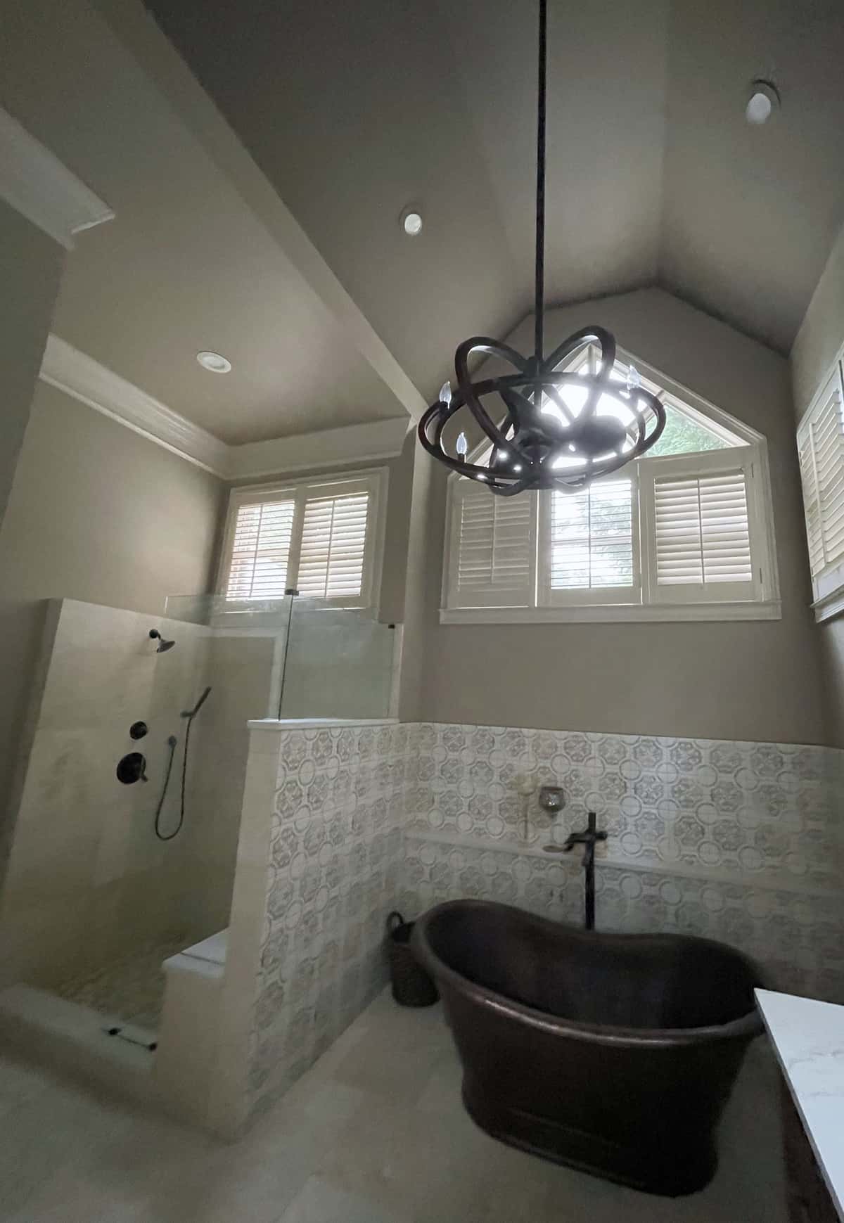 Hammered copper tub with a floor-mounted filler beneath an orb chandelier, beside a tiled walk-in shower
