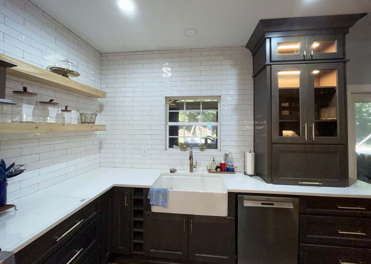 Espresso kitchen with a white farmhouse sink, floating oak shelves, glossy white subway tile and a lit glass-front cabinet tower