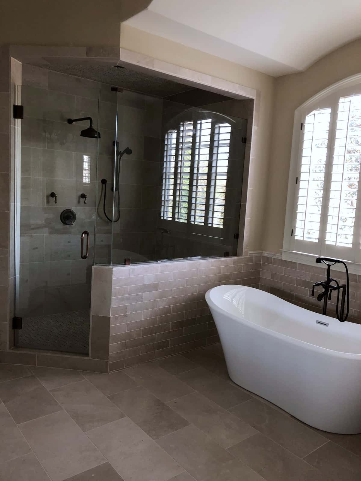 Frameless glass shower with a tiled half wall, freestanding tub and plantation shutters in a travertine-look primary bath