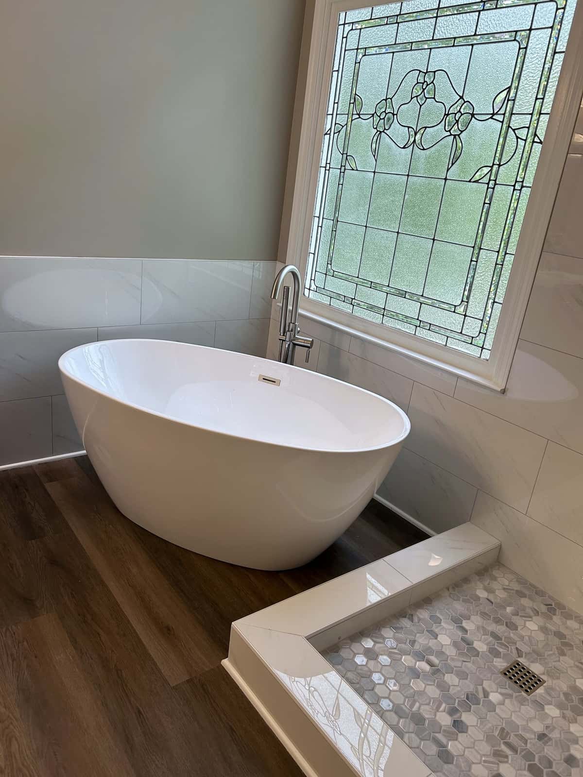 White freestanding tub with a floor-mounted filler beneath a leaded glass window, beside a hex mosaic shower threshold