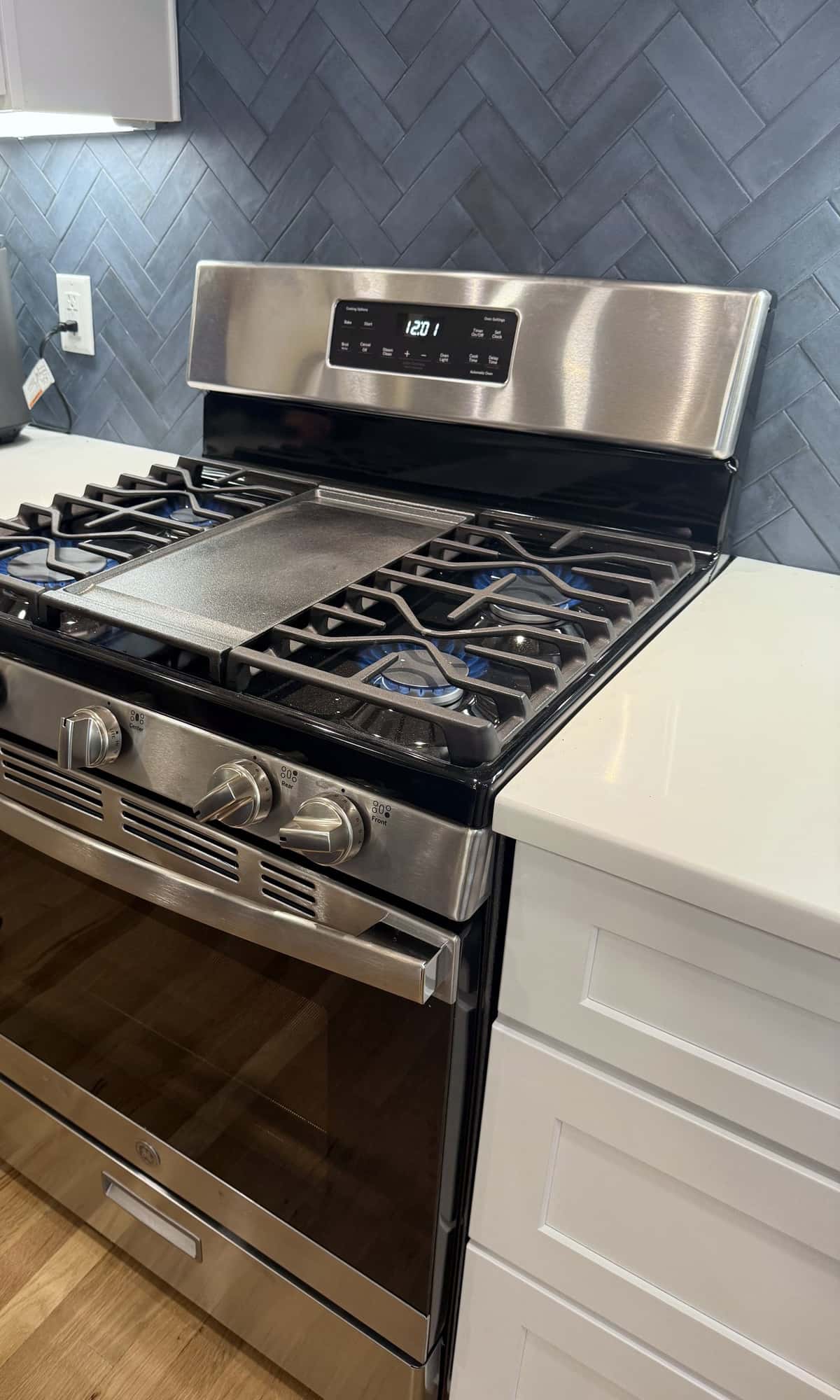 Stainless gas range set into white shaker cabinetry with a navy herringbone tile backsplash and white quartz