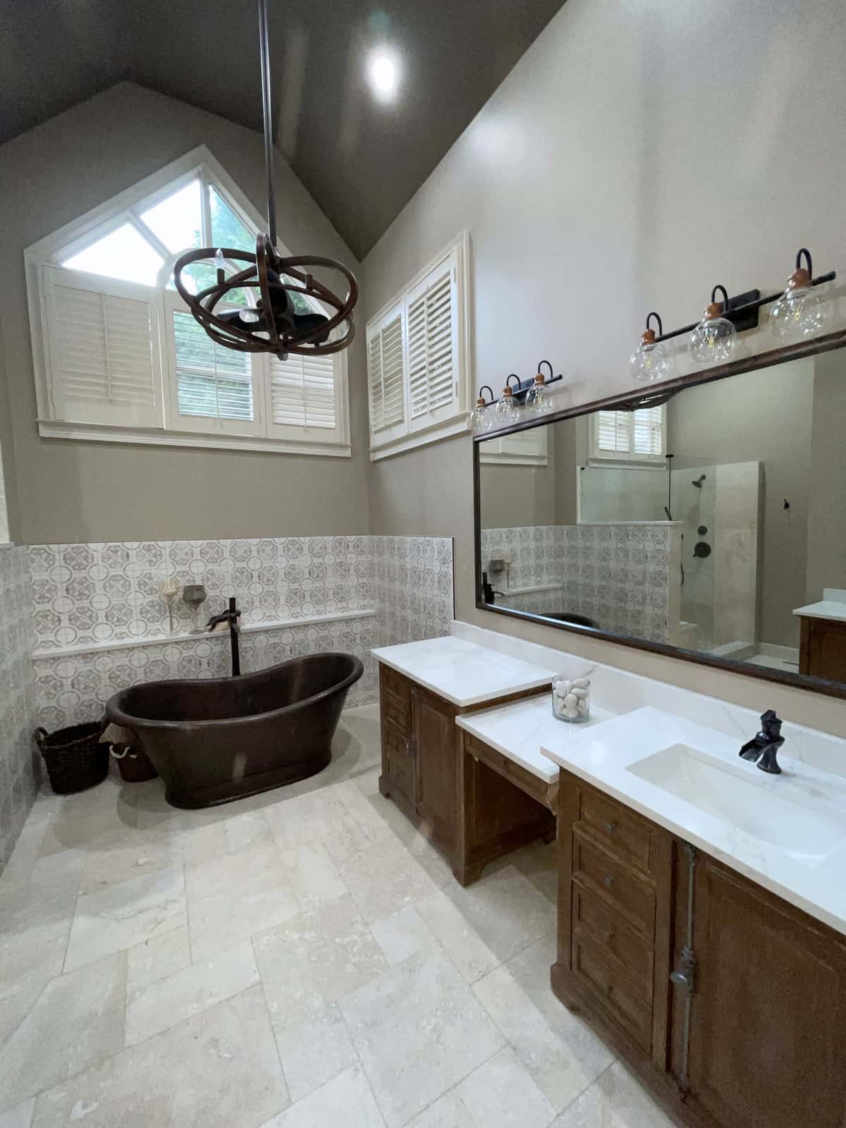 Vaulted primary bath with a hammered copper tub, patterned tile wainscot, wood double vanity and plantation shutters