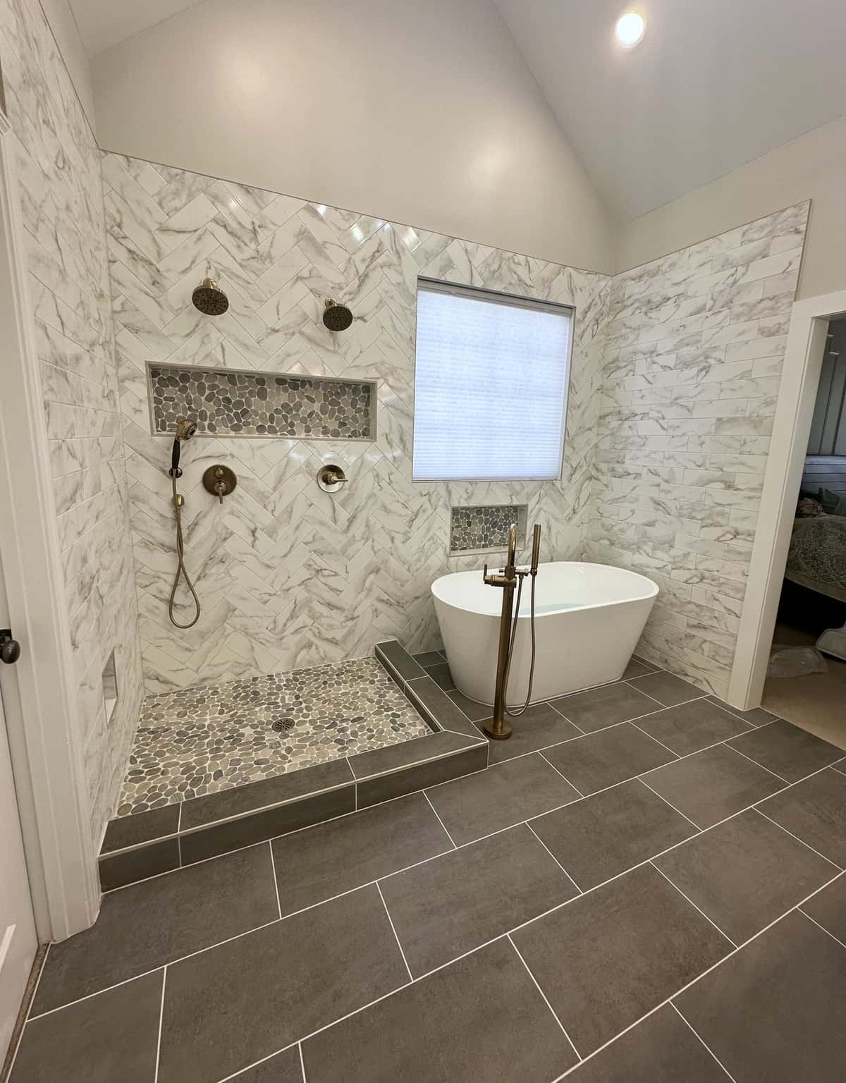 Vaulted wet room with herringbone marble-look tile walls, a pebble shower floor, brushed gold fixtures and a freestanding tub