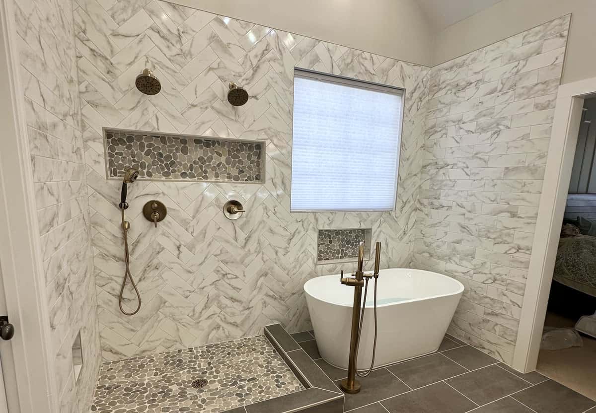 Vaulted wet room with herringbone marble-look tile, a pebble shower floor, brushed gold fixtures and a freestanding tub by Colitz Construction