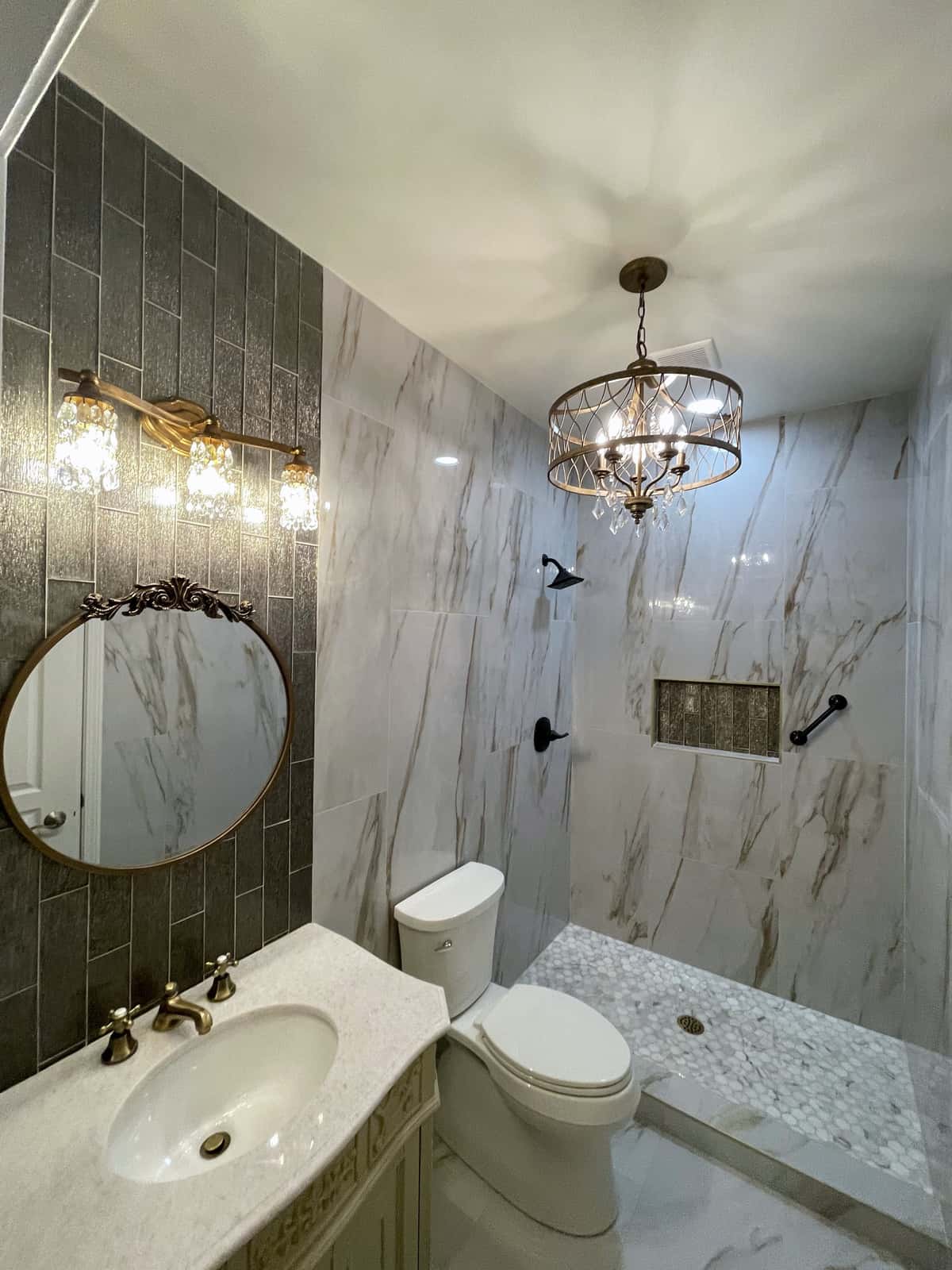 Bath with a marble-look tile shower, charcoal vertical tile accent wall, crystal chandelier, round gold mirror and brass fixtures