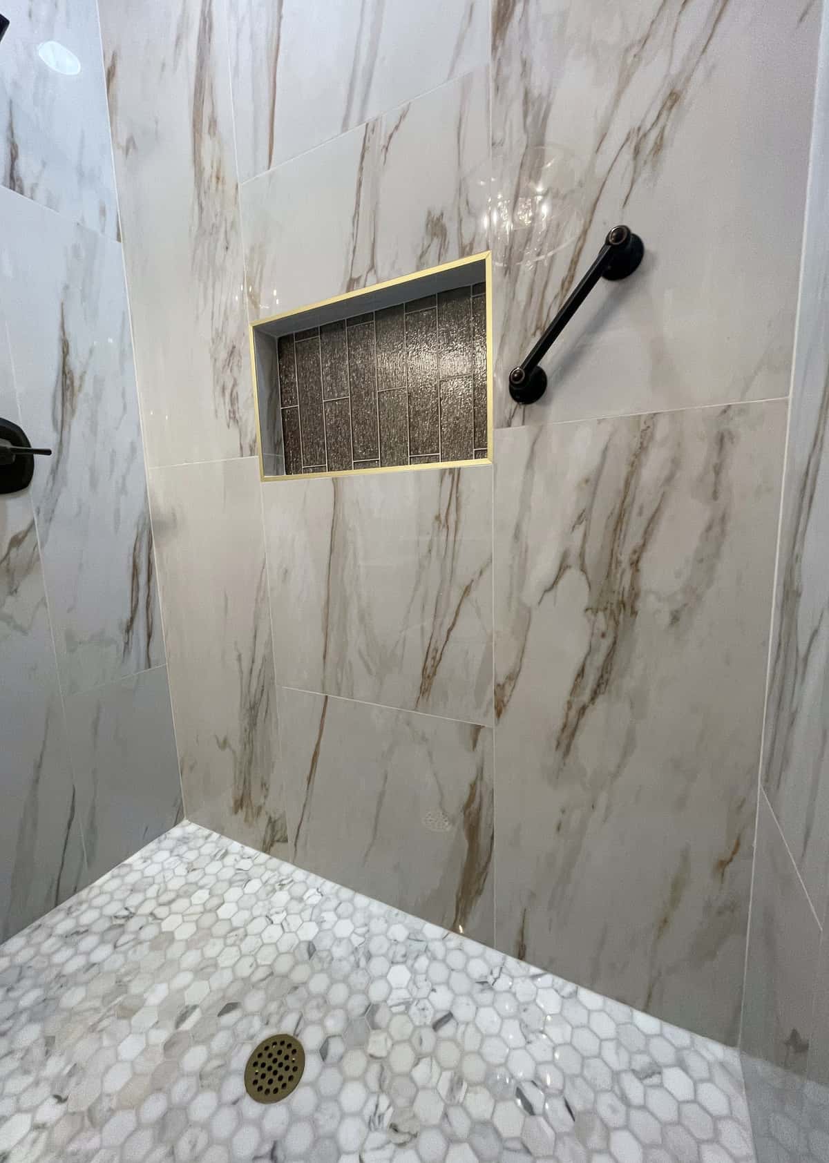 Curbless marble-look shower with a brass-framed charcoal tile niche, matte black grab bar and hex mosaic floor