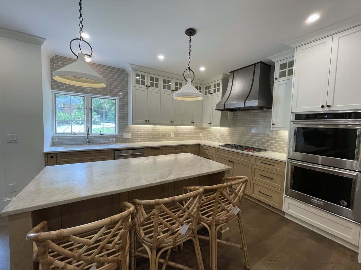 Taupe shaker kitchen with a veined stone island, rattan counter stools, white pendants, a black hood and double wall ovens