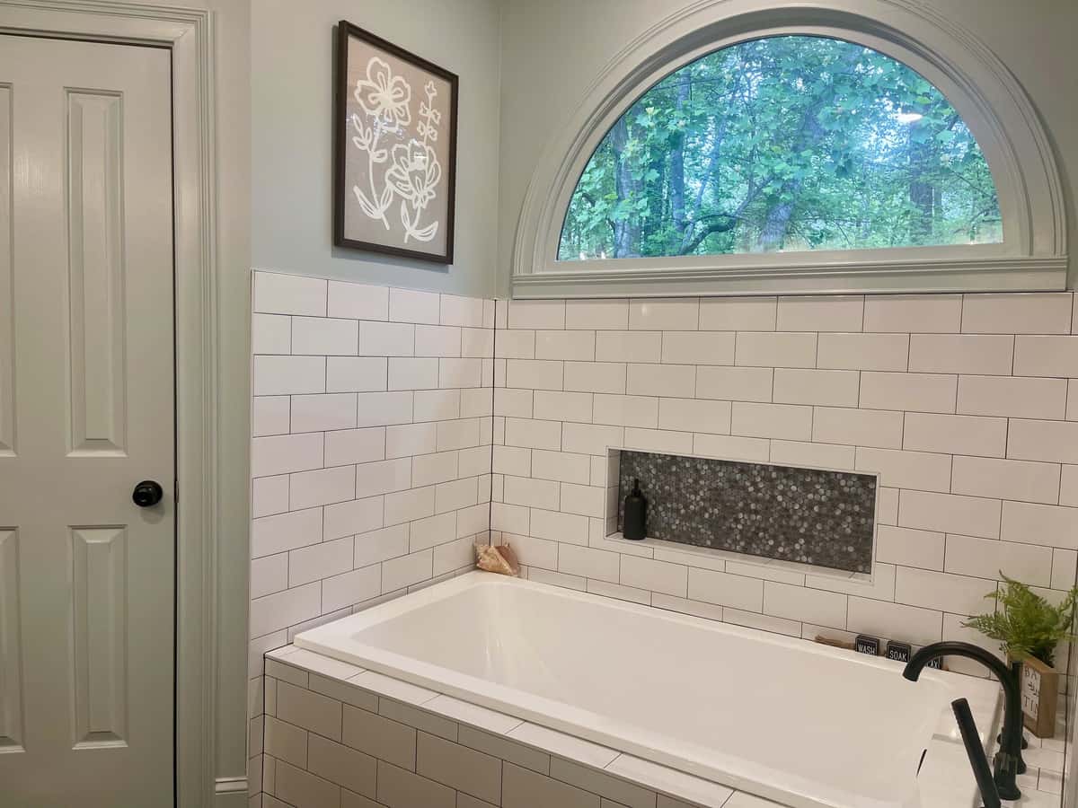 Drop-in soaking tub surrounded by white subway tile with a black pebble mosaic niche beneath an arched window