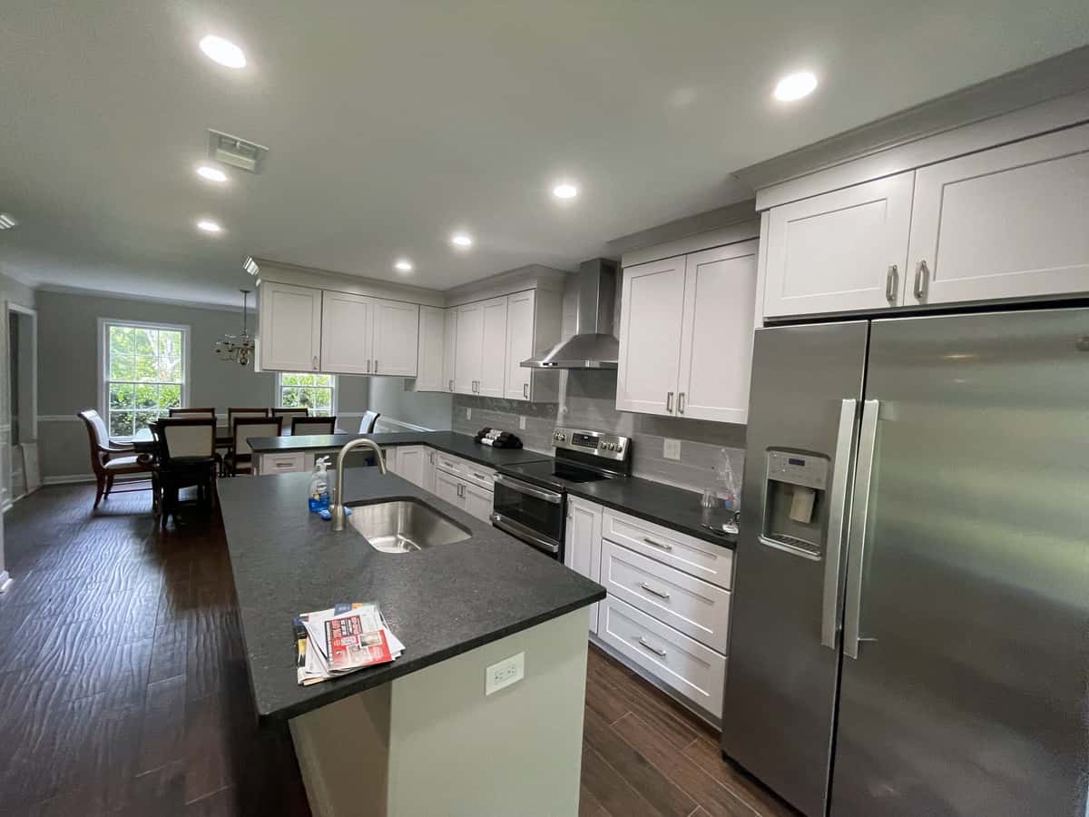 Open white shaker kitchen with a black granite island, stainless appliances and dark wood floors leading to the dining area