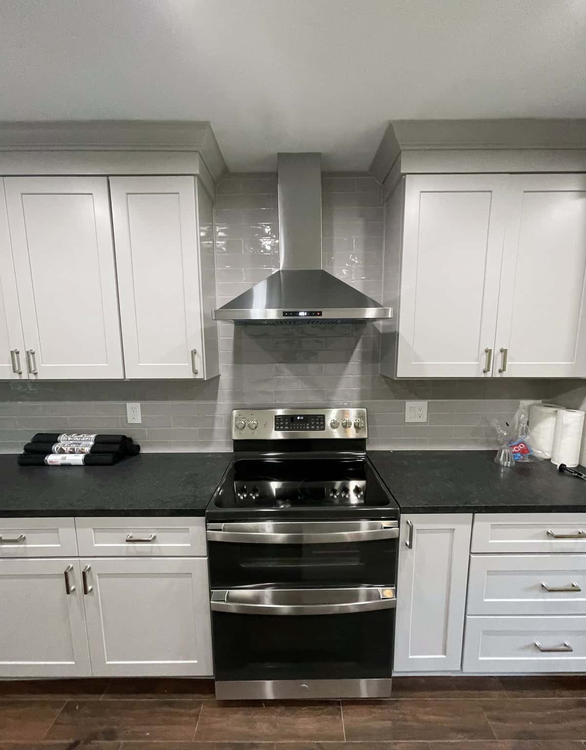 White shaker range wall with a stainless chimney hood, double-oven range, gray glass subway tile and black granite counters