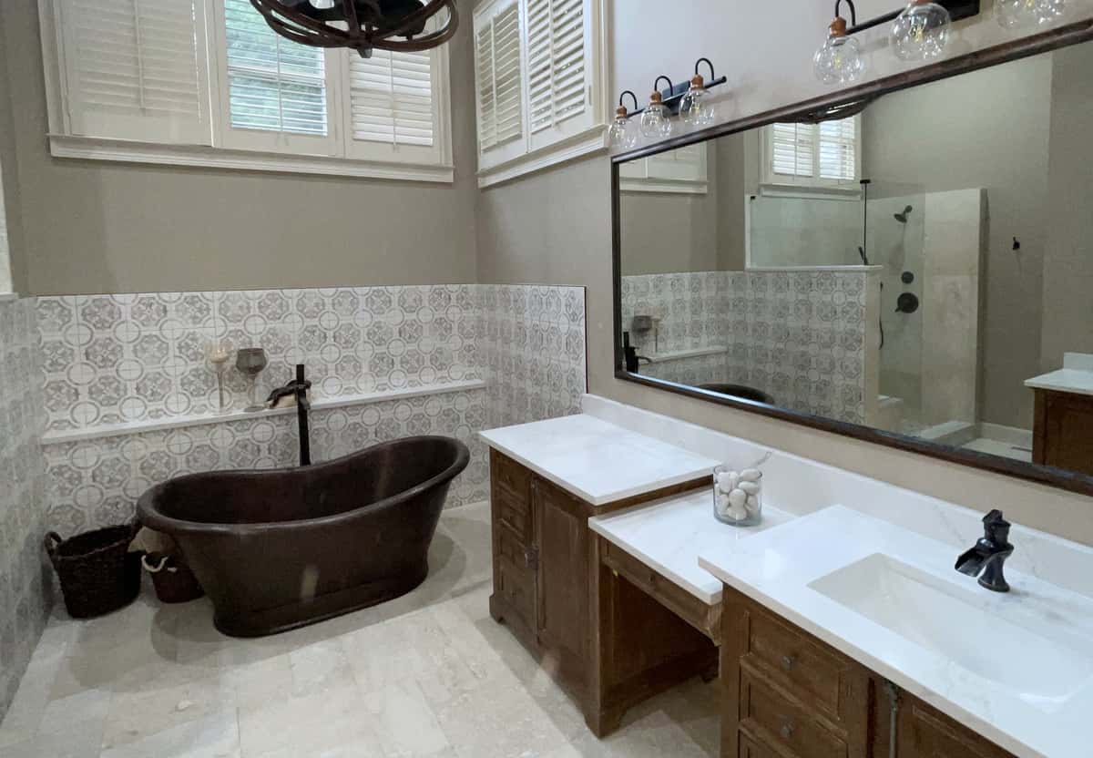 Vaulted primary bath with a hammered copper tub, patterned tile wainscot, wood double vanity and plantation shutters