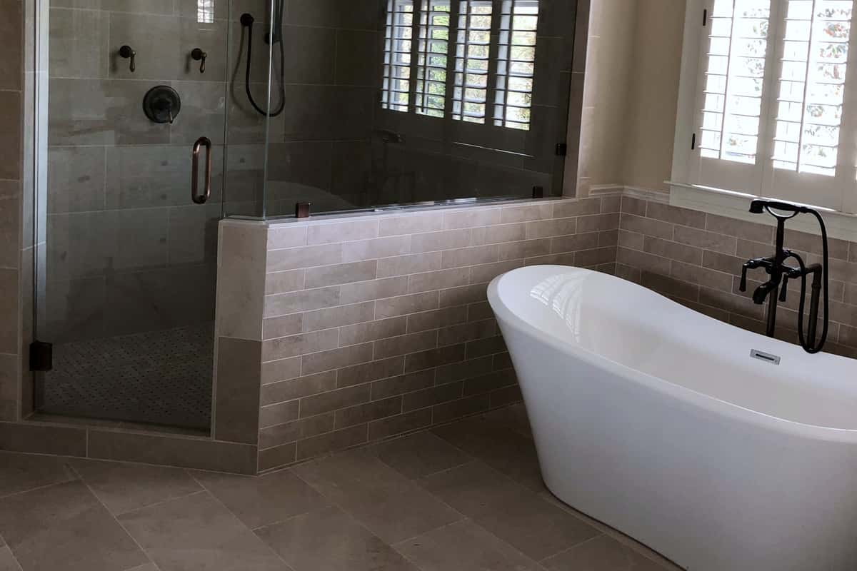 Frameless glass shower with a tiled half wall beside a freestanding tub and plantation shutters